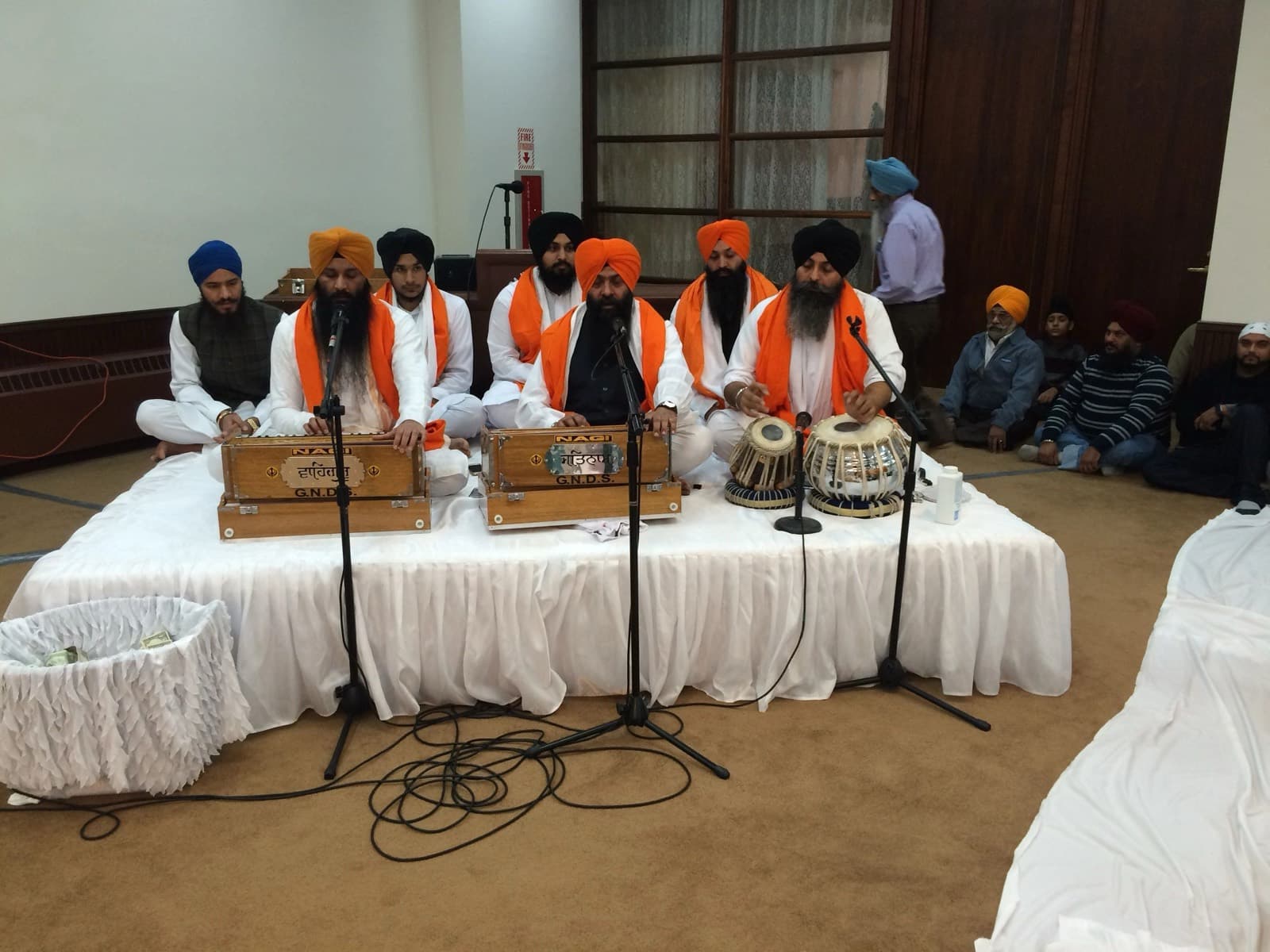 A kirtan jatha in white with saffron scarves on the Darbar Hall stage, two harmoniums marked G.N.D.S. and a tabla, sangat seated on the carpet