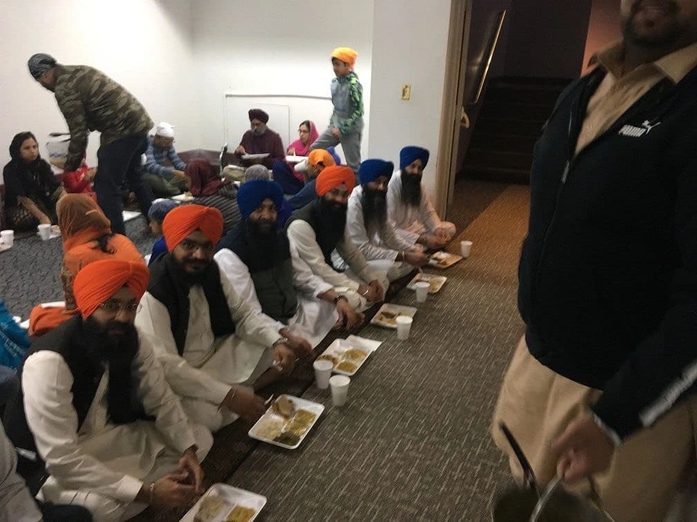 Sangat seated in rows on the Langar Hall floor eating langar from trays while sewadars serve