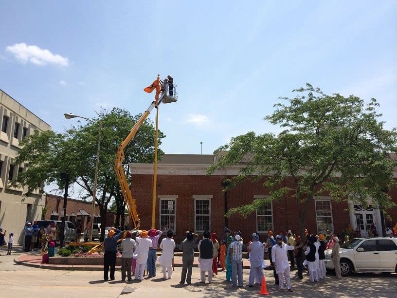 Sangat gathered on the sidewalk as two sewadars in a cherry-picker raise the saffron Nishan Sahib beside the red-brick gurdwara