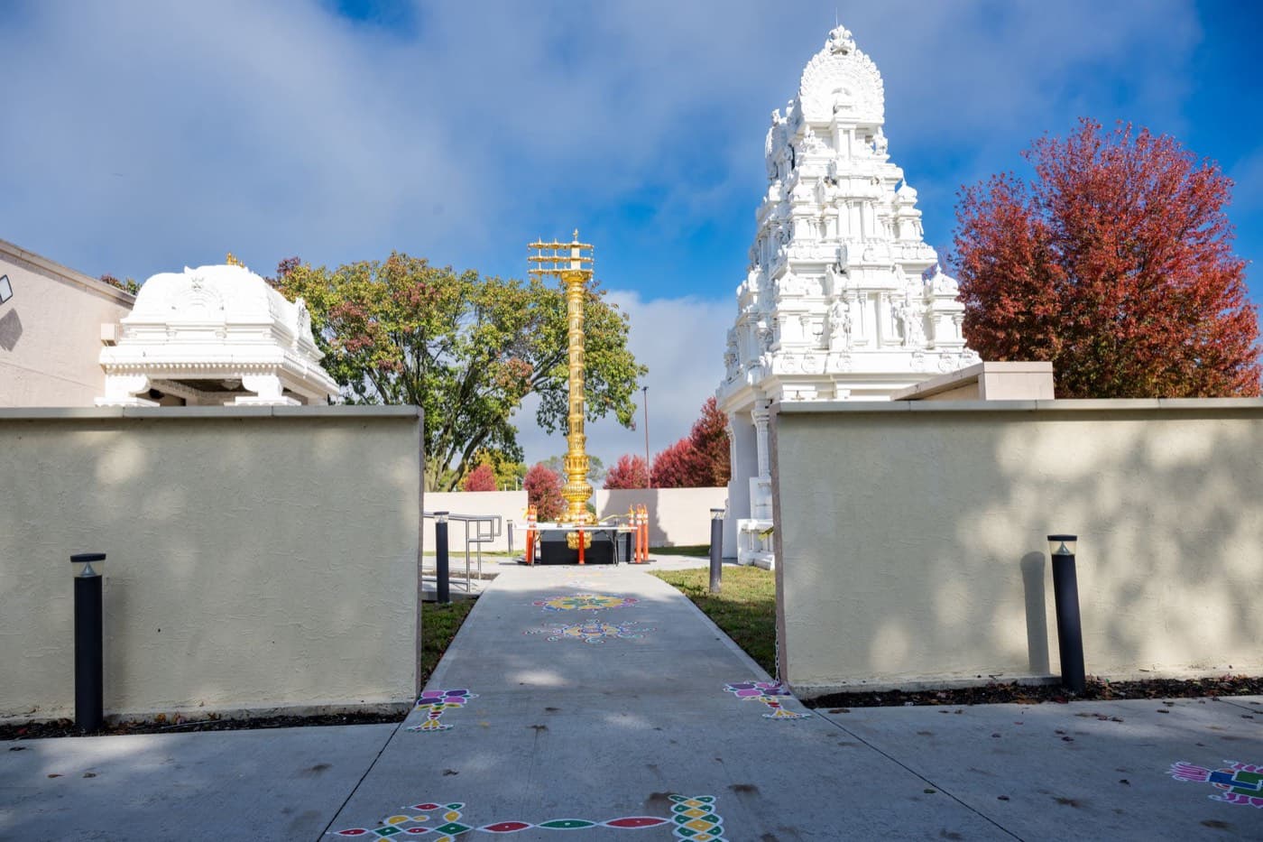 The gold-plated Dwajasthambam flagstaff standing between the temple's white Rajagopuram and the courtyard shrine, with rangoli on the walkway and autumn trees behind