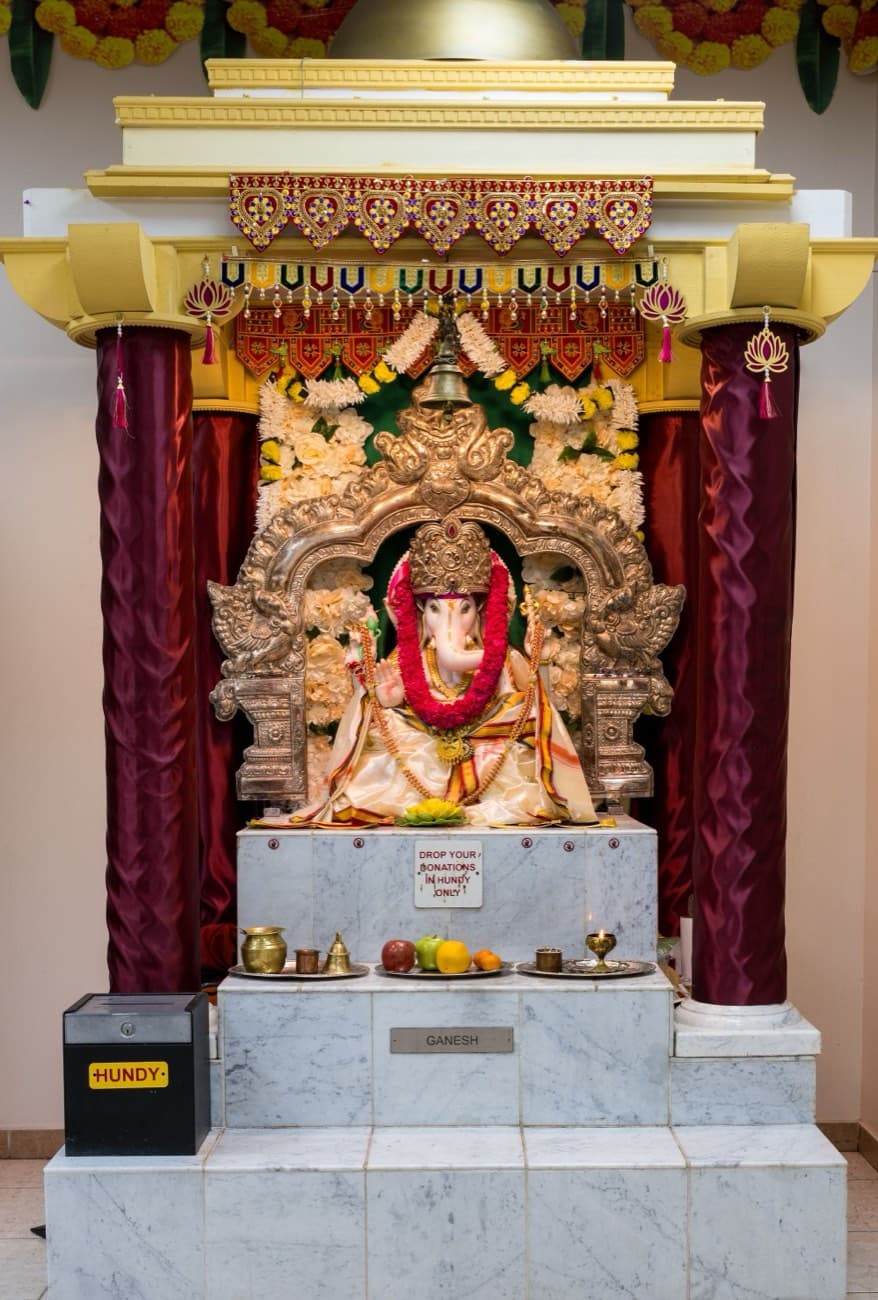 The Ganesh shrine at HTCC Kansas City, the marble Ganesha in a gold arch under a floral canopy with a hundi box beside it