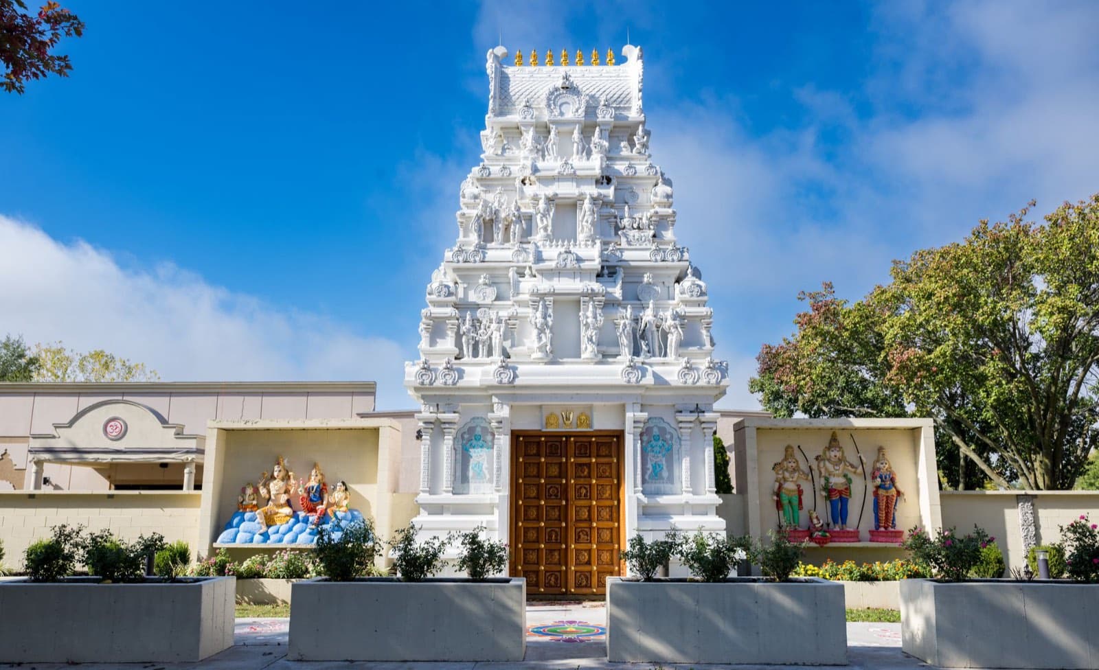The white three-tier Rajagopuram gateway of the Hindu Temple & Cultural Center of Kansas City on Lackman Road, Shawnee — carved wooden doors, painted deity reliefs on the flanking walls, blue sky above