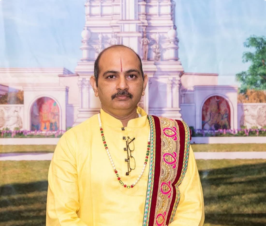 Priest Shree Pritam of HTCC Kansas City in a yellow kurta and embroidered shawl, in front of the temple backdrop