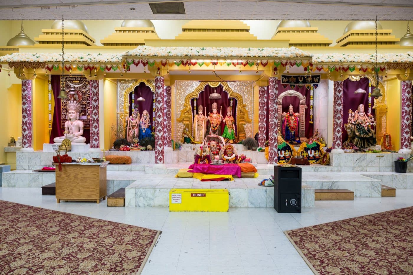 The main worship hall at HTCC Kansas City: a row of marble shrines under a red-and-gold mandap, with the central Sri Venkateswara altar and carpeted seating either side of the aisle