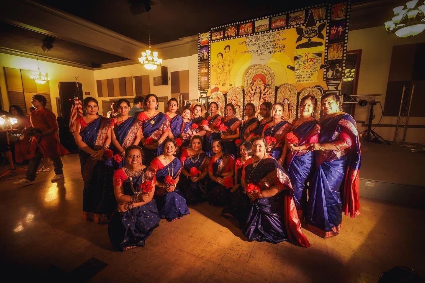 The KCBA ladies in matching blue-and-red saris holding red carnations after their Durga Puja 2022 dance, in front of the idols at the Scottish Rite