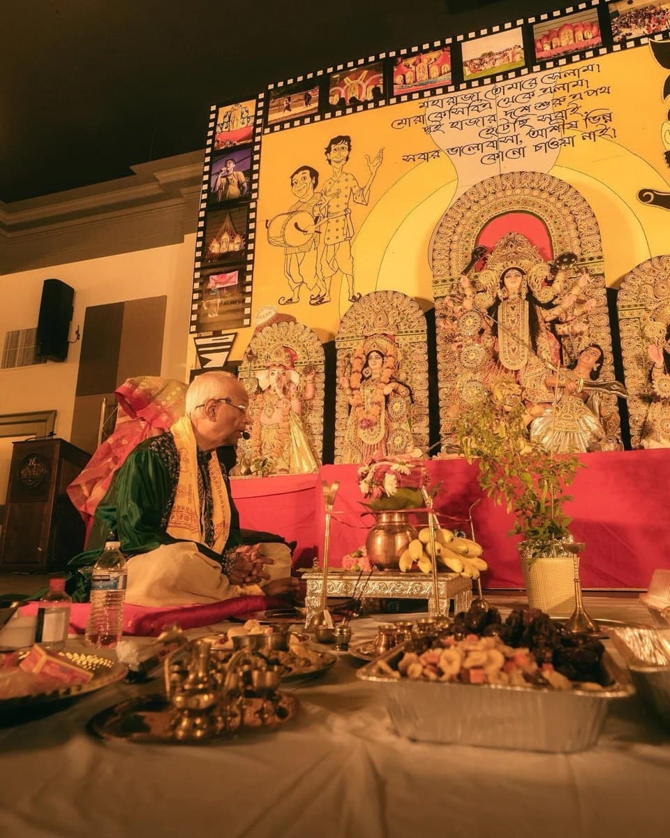 The priest seated before the fruit, flowers and brass lamps of the puja at KCBA's Durga Puja 2022, the idols and Gupi–Bagha backdrop behind him