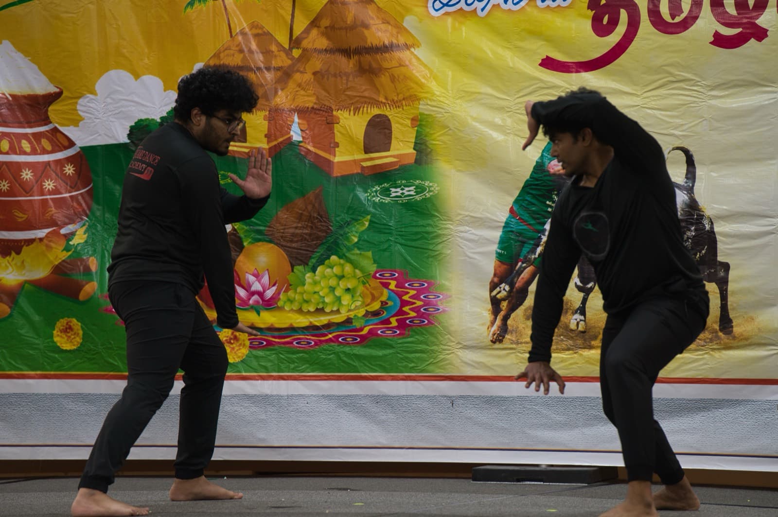 Two dancers mid-step on stage in front of the Pongal backdrop at Kansas City Tamil Sangam's Pongal 2024 celebration