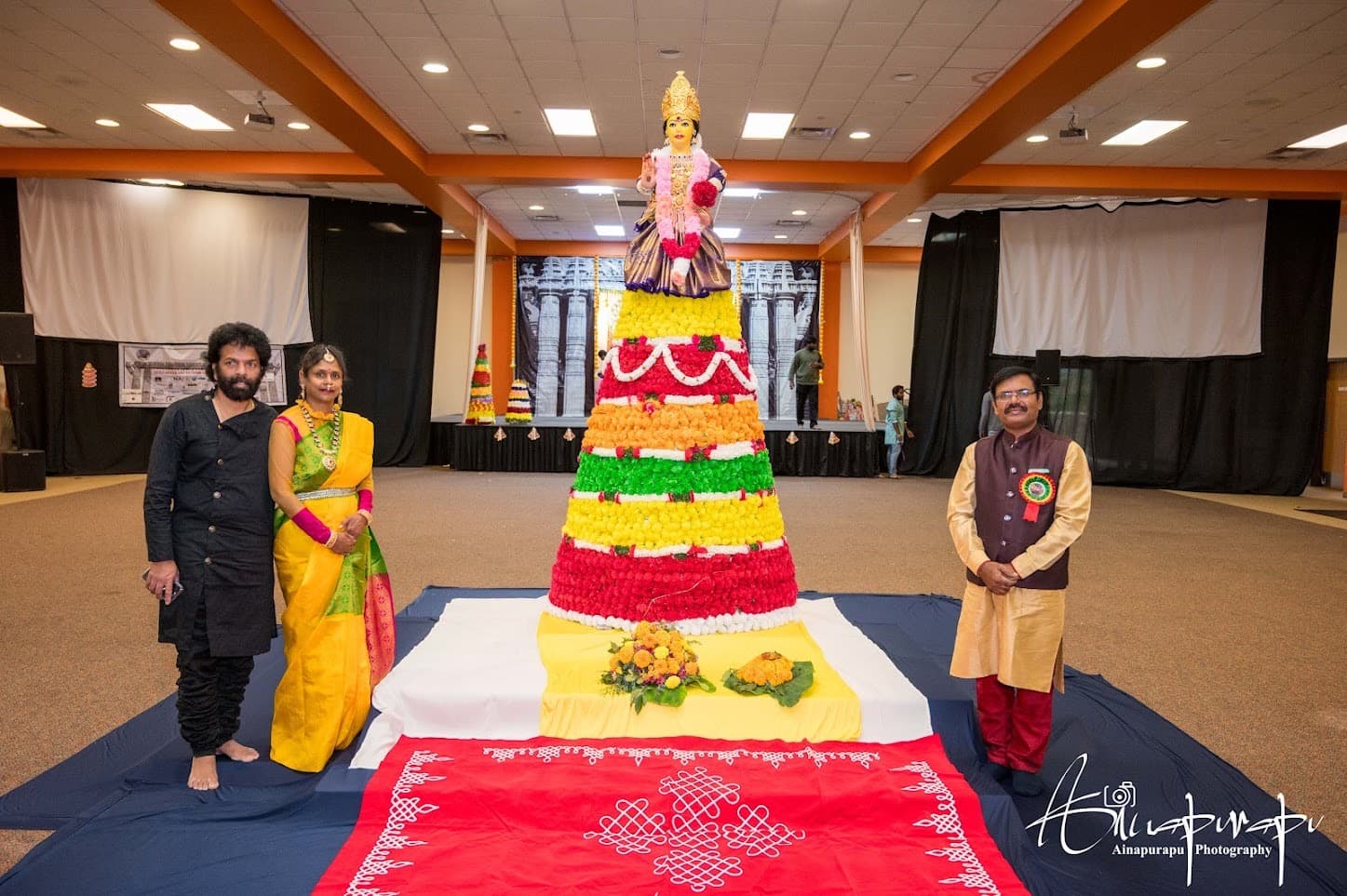 A person-height Bathukamma of red, orange, green and yellow flowers topped with a Gowramma idol on a rangoli cloth at KCTCA's Bathukamma 2023