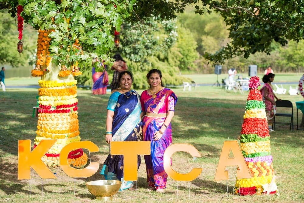Two women in silk sarees beside orange KCTCA letters, a marigold-wrapped tree and a tall flower Bathukamma at KCTCA's outdoor Bonalu celebration, 2023