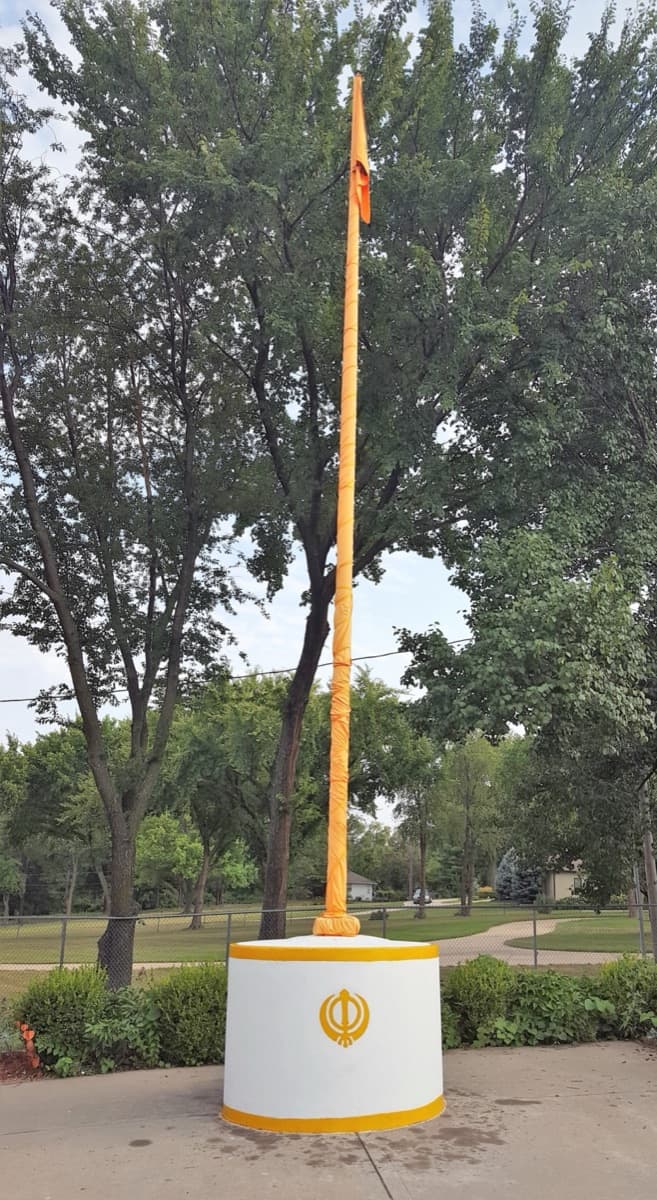 The Nishan Sahib — the saffron Sikh flag on its tall wrapped pole rising from a white base marked with the Khanda — outside the gurdwara